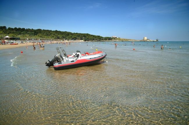Gommone rosso-nero nelle acque limpide, spiaggia e bagnanti sullo sfondo.
