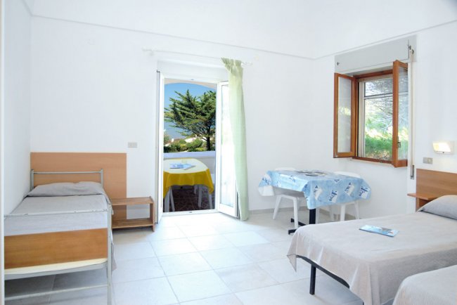Camera luminosa con due letti singoli, una porta-finestra aperta su un balcone con vista sul mare.
