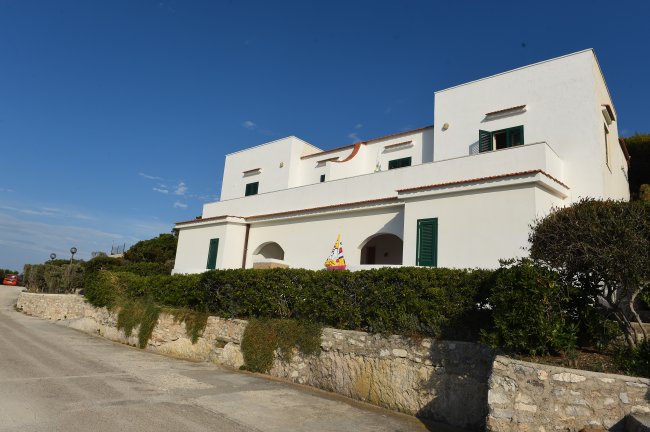 Edificio bianco moderno con finestre verdi, circondato da siepi e un muro di pietra.