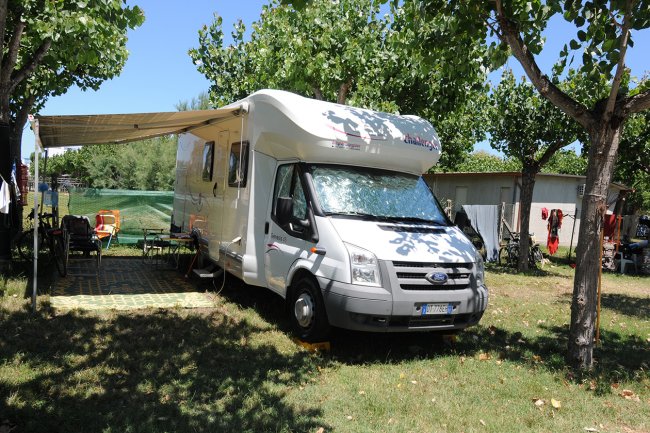 Camper bianco parcheggiato all'ombra di alberi, con sedie e tavolo sotto un telo esteso.