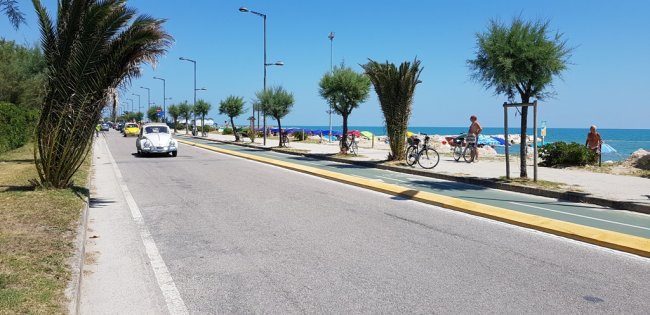 Strada costiera con una pista ciclabile, auto, biciclette e vista sul mare.