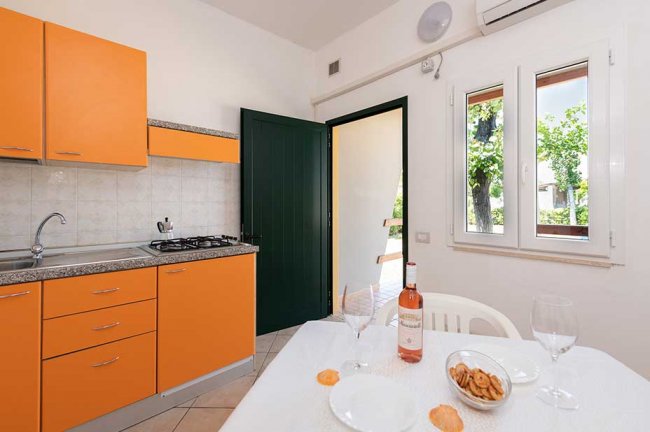 Cucina interna con mobili arancioni, tavolo apparecchiato e porta aperta su un balcone.