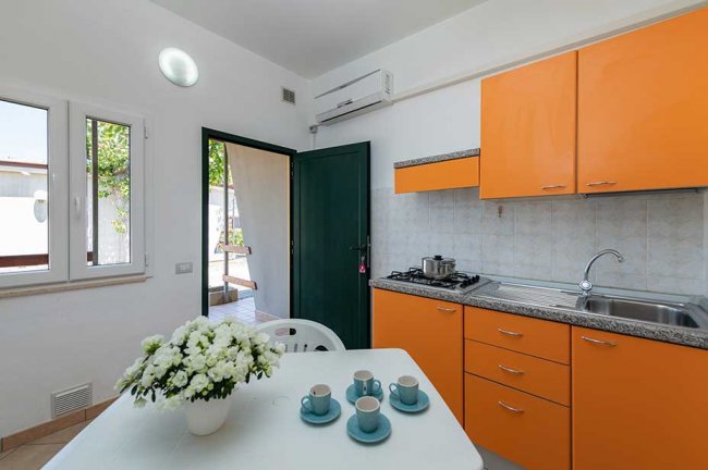 Cucina con mobili arancioni, tavolo bianco e fiori. Porta aperta verso l'esterno.