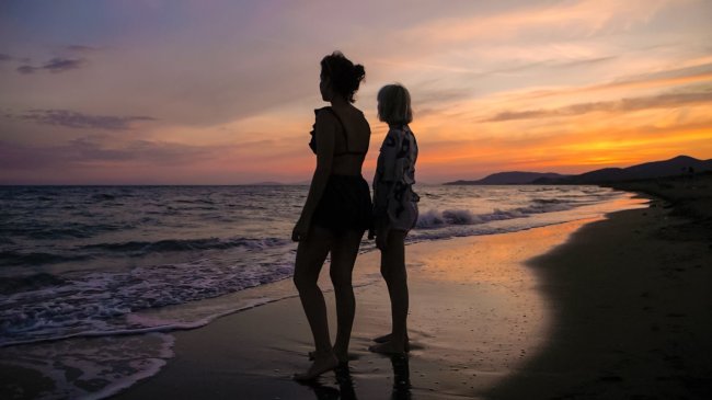 Due persone guardano il tramonto sulla spiaggia, illuminate dalla luce arancione del cielo.