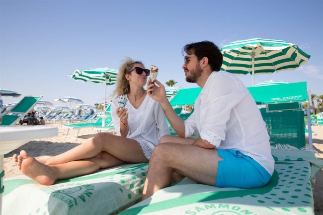 Due persone su una spiaggia, sedute su lettini, mangiano gelato sotto ombrelloni verdi e bianchi.