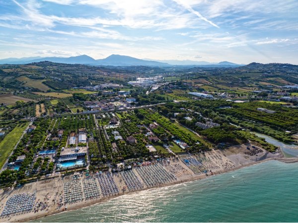 Vista aerea di una spiaggia affollata con ombrelloni e resort circondati da colline e vegetazione.