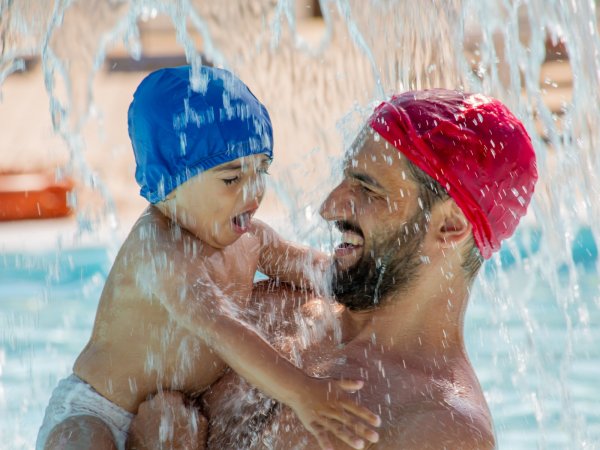 Uomo con cuffia rossa e bambino con cuffia blu giocano sotto una cascata in piscina.