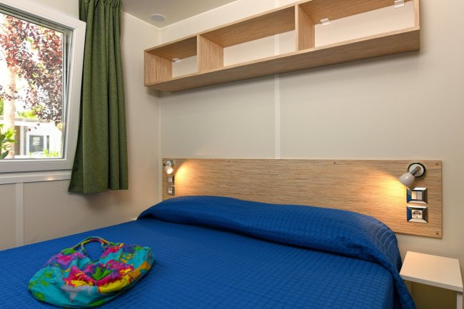 Camera con letto matrimoniale, copriletto blu, mensola e finestra con tenda verde.
