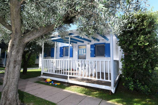 Casetta bianca con persiane blu e veranda, circondata da alberi.