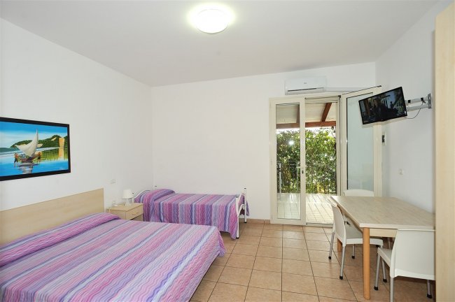 Camera con due letti singoli, arredi chiari, TV e porta finestra su veranda.