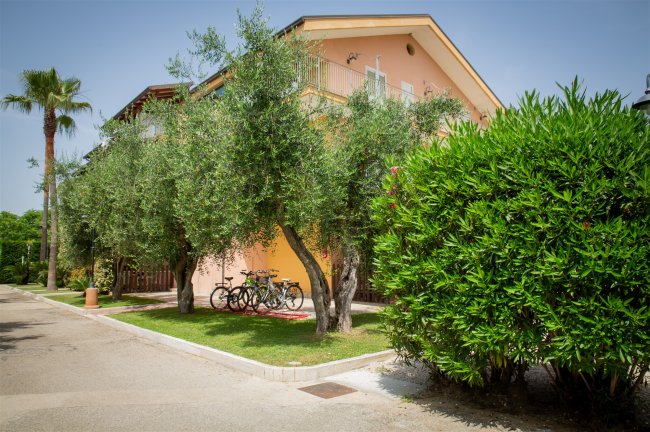 Esterno di edificio rosa con balconi, biciclette parcheggiate e alberi verdi intorno.