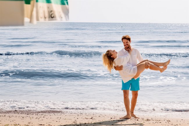 Un uomo solleva una donna sorridente su una spiaggia, davanti al mare calmo.