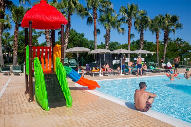 Piscina con scivolo colorato e persone rilassate sotto ombrelloni.