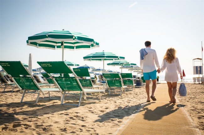Coppia cammina sulla spiaggia tra lettini e ombrelloni verdi e bianchi.