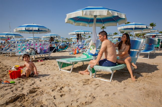 Famiglia in spiaggia sotto ombrelloni blu e bianchi.