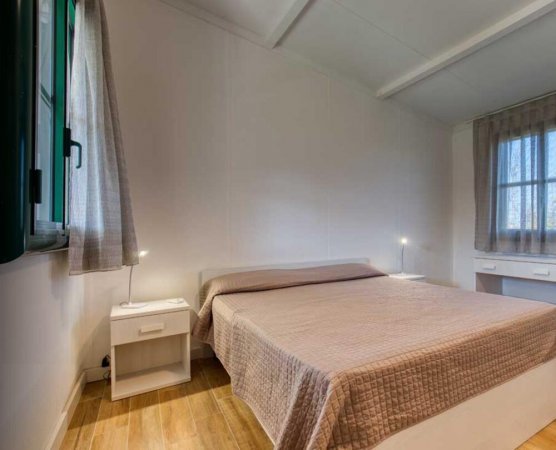 Un letto matrimoniale con copriletto beige, due comodini bianchi e due finestre con tende.