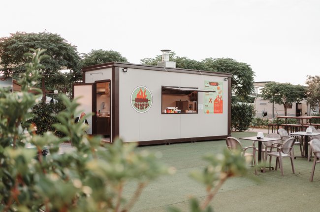 Un piccolo chiosco con un'insegna di hamburger, circondato da alberi e sedie all'aperto.
