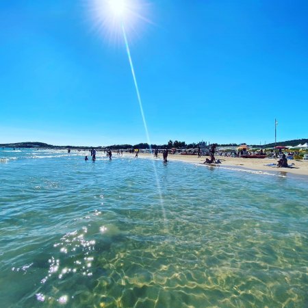 Spiaggia soleggiata con persone in acqua e sole brillante nel cielo azzurro.