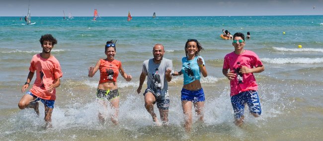 Cinque persone sorridenti corrono in mare, con il cielo azzurro sopra e vele sullo sfondo.