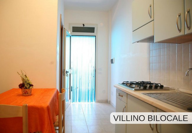 Cucina con tavolo arancione e ingresso con porta trasparente. Scritta: 
