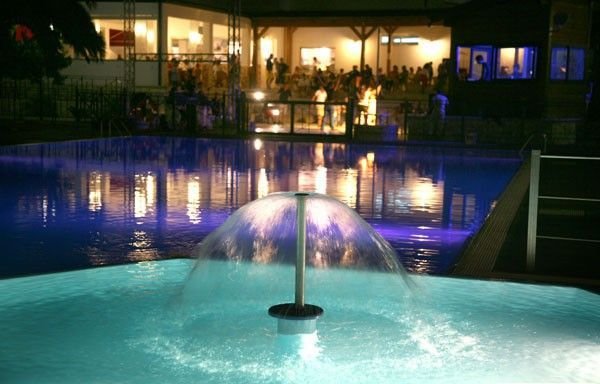 Piscina notturna illuminata con fontana al centro.
