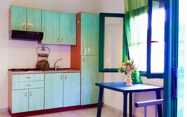 Cucina con pensili verde acqua, tavolino blu con vaso di fiori e finestra con tenda verde.