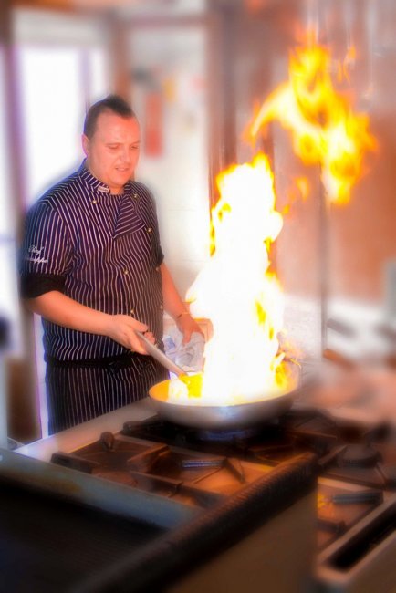 Uno chef in divisa cucina con una padella da cui si sprigiona una grande fiamma.