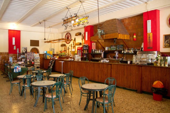 Interno di un bar con sedie e tavoli, un bancone lungo e decorazioni rustiche.
