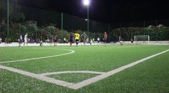 Campo da calcio notturno illuminato, con giocatori in azione e una rete sullo sfondo.