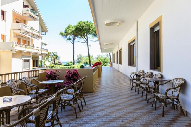 Una terrazza con tavoli e sedie, palme e vista su alberi verdi e cielo azzurro.
