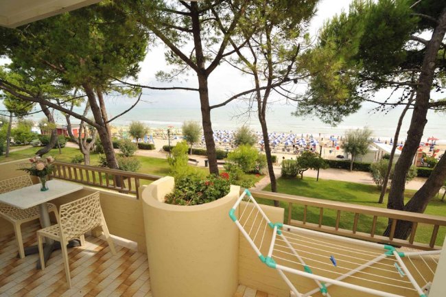 Un balcone con tavolino, sedie, stendino e vista mare, circondato da pini.