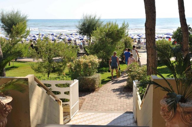 Scalinata verso una spiaggia con persone e ombrelloni blu bianchi, alberi e cespugli.