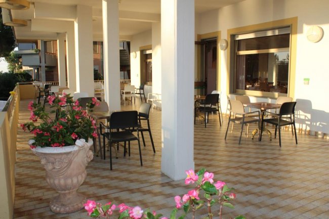 Terrazza coperta con colonne bianche, tavoli, sedie e vasi di fiori.