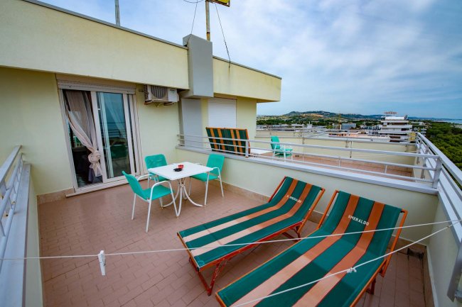 Terrazza con due sdraio a righe, un tavolo bianco e sedie verdi. Visuale della città.