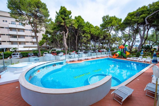 Piscina circondata da alberi e sedie a sdraio, con edifici sullo sfondo.