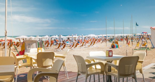 Tavoli e sedie di plastica su una terrazza con vista su una spiaggia con ombrelloni.