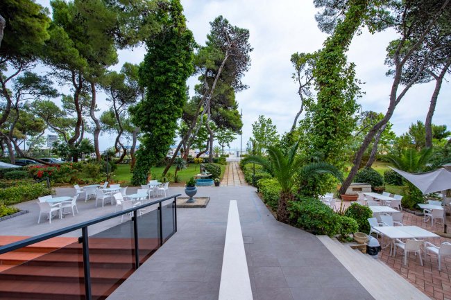 Sentiero fiancheggiato da alberi e vegetazione lussureggiante, con tavoli all'aperto.