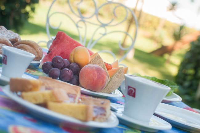 Tavolo da colazione all'aperto con frutta, dolci e tazze di caffè su una tovaglia colorata.