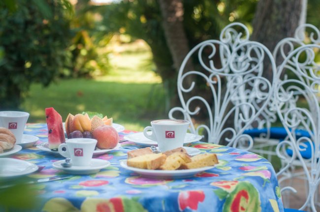 Tavolino all'aperto apparecchiato con colazione, frutta e caffè, sedie bianche decorative intorno.