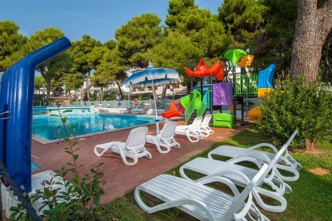 Area piscina con sdraio e ombrelloni, vicino a un parco giochi per bambini con scivoli colorati.