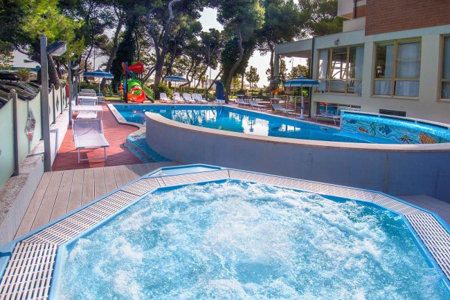 Area esterna con piscina e vasca idromassaggio circondate da lettini e alberi.
