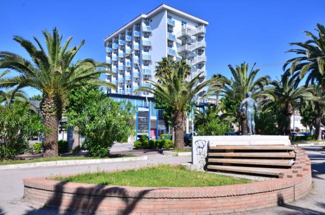 Alta palazzina moderna con balconi, circondata da palme e un'aiuola con una statua.