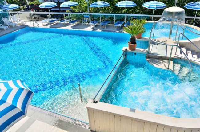 Piscina azzurra con ombrelloni a righe blu e bianche, vasca idromassaggio e piante in vaso.