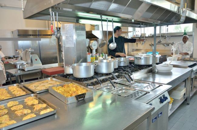 Cucina industriale con pentole in acciaio, teglie di cibo e due persone al lavoro.