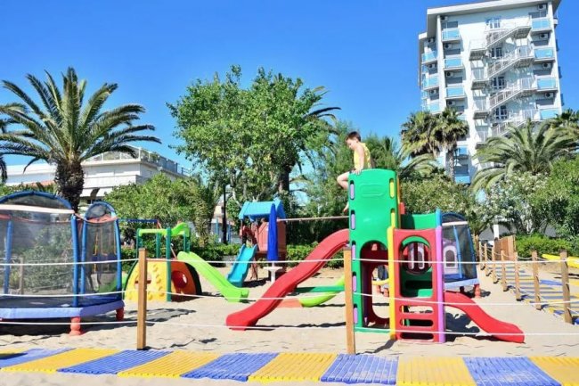 Parco giochi con scivoli colorati su sabbia, palme e un edificio sullo sfondo.