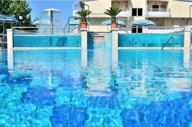 Piscina blu con cascate e ombrelloni, edificio bianco sullo sfondo.