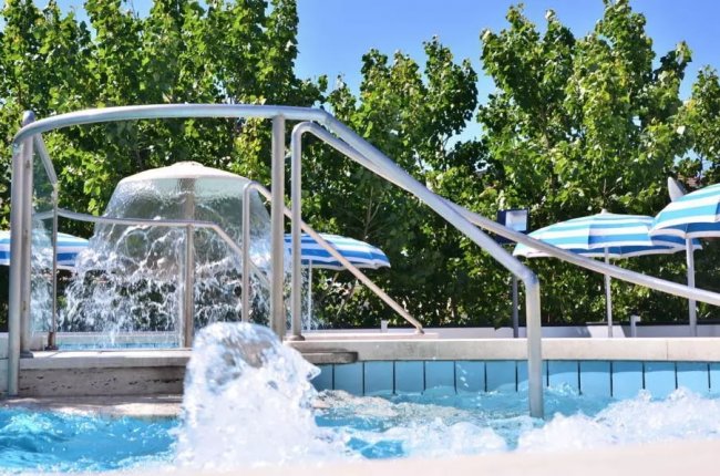 Una piscina con scivolo, giochi d’acqua e ombrelloni a righe blu, circondata da alberi verdi.