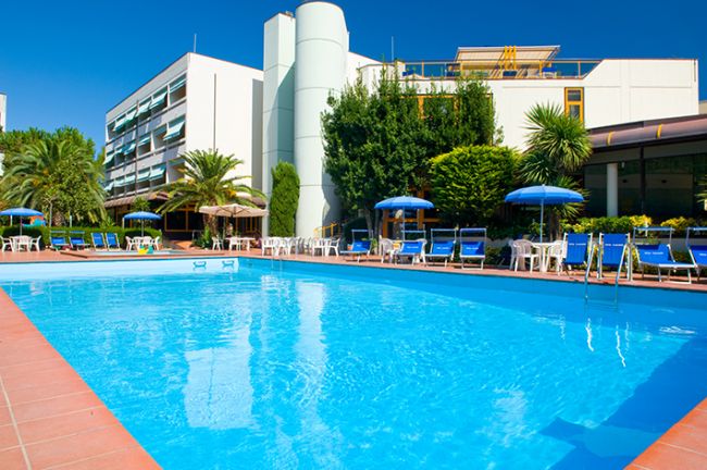 Piscina azzurra circondata da sedie a sdraio blu e palme, con un grande edificio sullo sfondo.