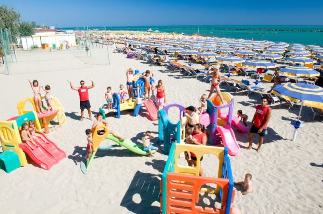 Bambini giocano su una spiaggia con scivoli colorati, mare in lontananza.