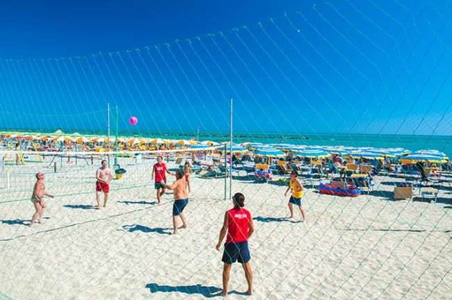 Persone giocano a beach volley su sabbia bianca, mare con ombrelloni gialli sullo sfondo.